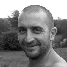 black and white photograph of a man with no shirt on smiling at the camera while standing in a field