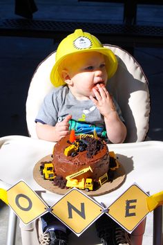 a baby wearing a yellow hat sitting in a chair with a construction cake on it