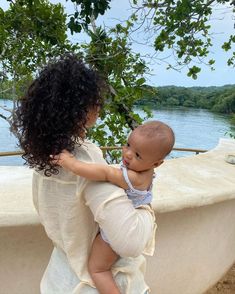 a woman holding a baby in her arms next to a body of water and trees