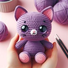 a hand holding a small purple crocheted cat next to yarn balls and knitting needles