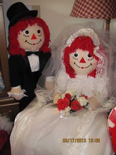 two stuffed dolls dressed as bride and groom sitting on a bed with red roses in the foreground