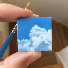 a person holding a small piece of paper with clouds on it and a paintbrush in their hand