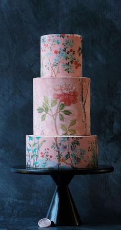 a three tiered cake decorated with flowers and leaves on a black stand against a blue background