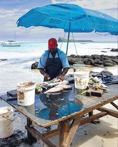 a man sitting at a table in front of an umbrella preparing food on the beach