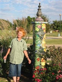 an older woman standing in front of a colorful pole with flowers on it and grass behind her