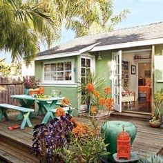 a small green house with flowers and potted plants on the front porch, next to a picnic table