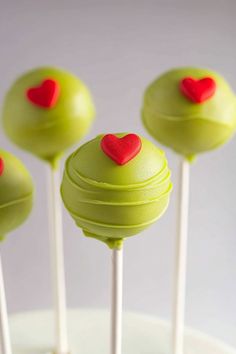 four green cake pops with red hearts on them sitting on top of a white plate