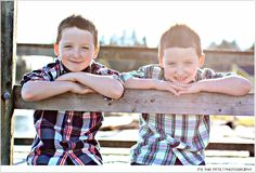 two young boys leaning on a wooden fence with their arms crossed and smiling at the camera