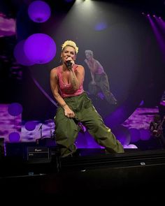 a woman on stage singing into a microphone with purple lights behind her and two other people in the background