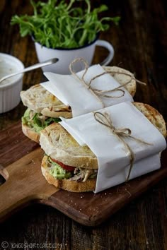 two sandwiches wrapped in white paper and tied with twine on a cutting board next to a bowl of greens