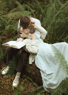 a man and woman are sitting on a log in the woods, reading a book