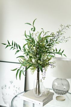 a glass table topped with a vase filled with greenery next to a white lamp