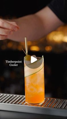 a tall glass filled with an orange drink on top of a metal table next to a person's hand