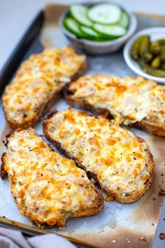 several pieces of bread with cheese and pickles on the side, sitting on a baking sheet