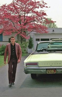 a man standing next to a green car in front of a tree with pink flowers