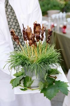 a man is holding a potted plant with meat on sticks in it and green leaves