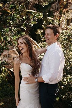 a man and woman standing next to each other in front of some bushes smiling at the camera