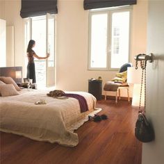 a woman standing next to a bed in a room with wooden floors and windows on both sides