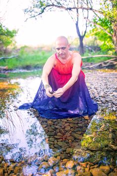 a man sitting on top of a rock covered river next to a small stream wearing a purple dress