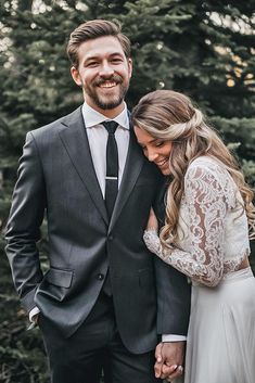 a bride and groom smile at each other as they stand in front of some trees