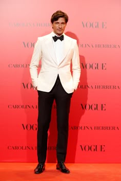 a man in a white tuxedo and black bow tie standing next to a red wall