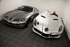 two silver sports cars parked next to each other in a white walled room with black tile flooring