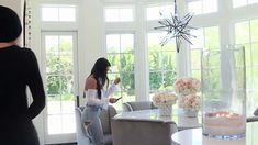 a woman standing in front of a living room window next to a table with flowers on it