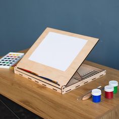 an easel with paint and pencils on it next to a wooden box that has a blank paper in it