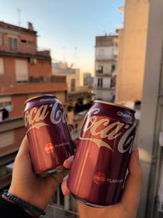 two cans of coca - cola are held up in front of the cityscape