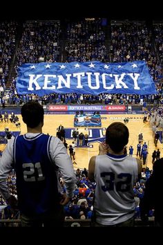 two men standing in front of a kentucky flag at a basketball game with people watching