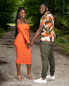 a man and woman holding hands while standing on a dirt road in the woods with trees behind them