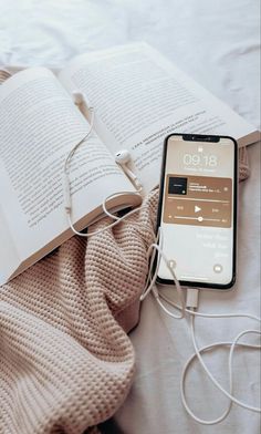 an open book and headphones are laying on a bed next to an iphone that is plugged in