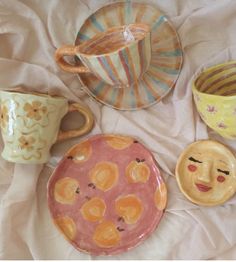 three ceramic plates and two cups on a bed with white sheets, one has a face painted on it