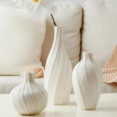 three white vases sitting on top of a table next to pillows and a couch