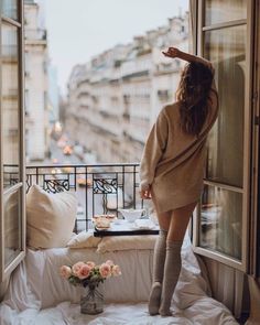 a woman standing on top of a bed in front of a window next to a vase with flowers