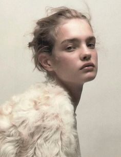 a woman with long hair wearing a fur coat and looking off to the side, in front of a white wall