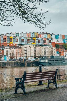 an empty park bench sitting next to a body of water with buildings in the background