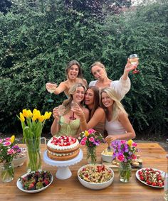 four women standing around a table with food and drinks in front of them, posing for the camera