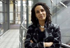 a woman with dreadlocks standing in front of a glass wall and stairs, looking at the camera