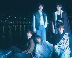 group of young men sitting on the ground in front of water at night with city lights behind them