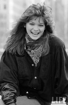 a black and white photo of a woman with long hair smiling at the camera while sitting on a bench