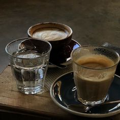 two cups of coffee sit next to each other on a table with spoons and glasses