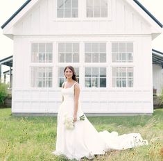 a woman standing in front of a white house with flowers on her wedding dress and bouquet