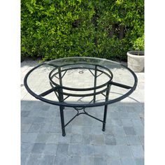 a glass table sitting on top of a stone floor next to green bushes and shrubbery