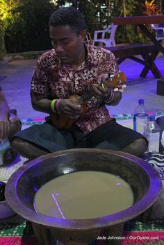 a man sitting on the ground playing a guitar and singing into a bowl of liquid