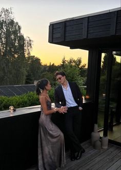 a man and woman standing next to each other on a wooden deck at sunset with trees in the background