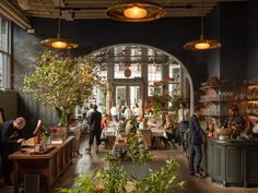 people are sitting at tables in a restaurant with potted plants and hanging lights above them