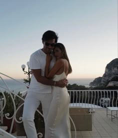 a man and woman embracing each other on a balcony overlooking the ocean in white outfits