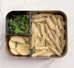 a metal container filled with pasta, broccoli and apple slices on top of a white counter