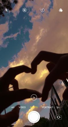 two hands making a heart shape in front of the sky with clouds and buildings behind them